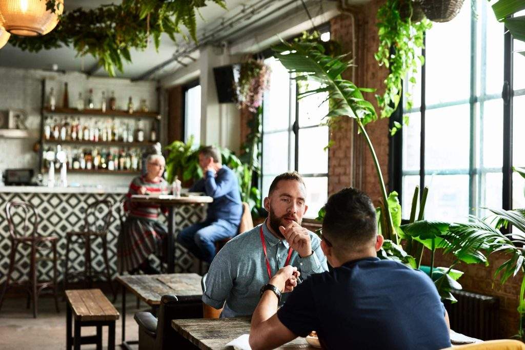 A manager interviews a candidate at a restaurant, with plants and a bar counter in the background