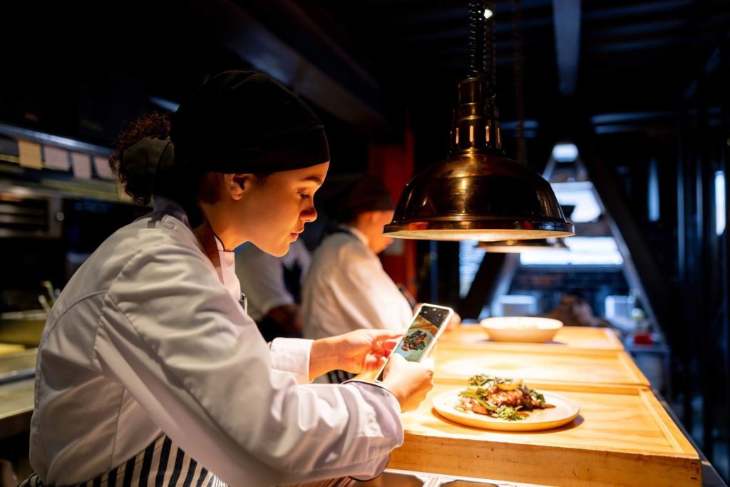 A restaurant chef is taking a picture of a dish they have prepared. They are using a mobile phone to share the picture on social media